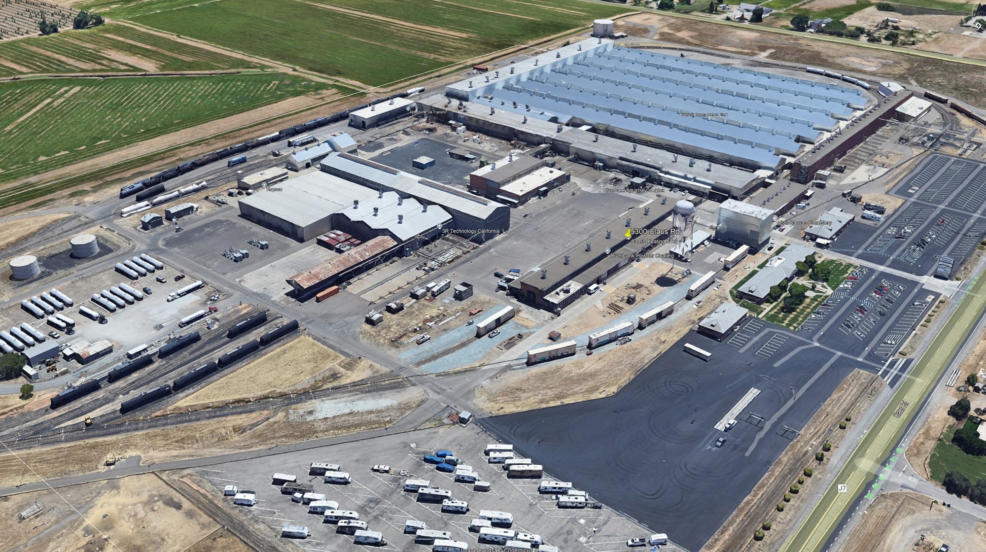 Aerial view of the Riverbank Industrial Complex: long industrial buildings, rail lines, and parking, with farmland beyond